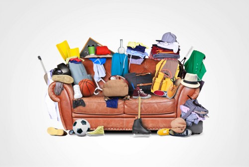 A brown fabric sofa heavily cluttered with a variety of discarded items, including sports equipment such as a football and basketball, clothing and textiles in different colours and fabrics, a tennis racket, a pair of shoes, and a hat. The sofa also holds office supplies like a notebook, and miscellaneous household objects. Surrounding the sofa on the white background are additional objects such as a yellow plastic container, a small chalkboard, and a toy car. The scene appears to depict a collection of rubbish prepared for removal, reflecting a typical cluttered environment in need of clearance services. The setting is plain with no visible environment, emphasizing the pile of assorted waste materials ready for rubbish collection, indicative of a typical domestic or office clearance scenario, possibly within or associated with Edmonton or nearby areas from the context of Office Clearance Edmonton's services.