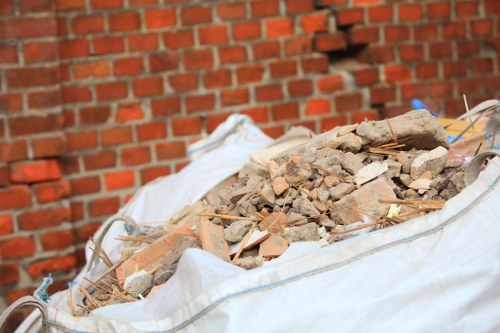 A large outdoor bag filled with a mixture of rubble and construction debris, including broken bricks, concrete fragments, and wooden pieces, situated on a paved surface against a red brick wall. The bag is made of white woven material and appears to be used for rubbish removal, with some of the debris spilling slightly over the edges. In the background, the brick wall extends horizontally, with visible mortar joints. The environment suggests a setting where building waste is being collected before disposal, consistent with rubbish removal services offered by Office Clearance Edmonton in the Edmonton area.