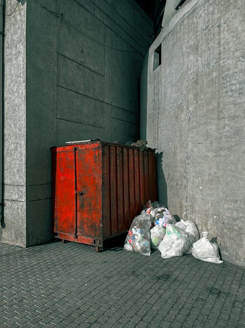 The image features a large, light red metal skip situated outdoors on a concrete surface near a white wall. The skip has a slightly weathered appearance with visible scratches and marks, and it is pos