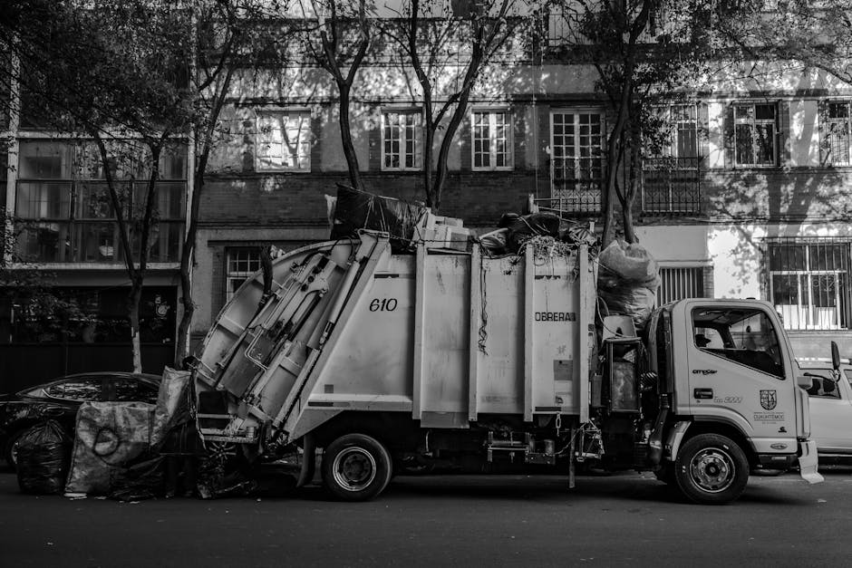 A worker dressed in a green uniform and safety helmet stands at the rear of a small waste collection vehicle, which is parked on a suburban street lined with trees and parked cars. The vehicle's rear 