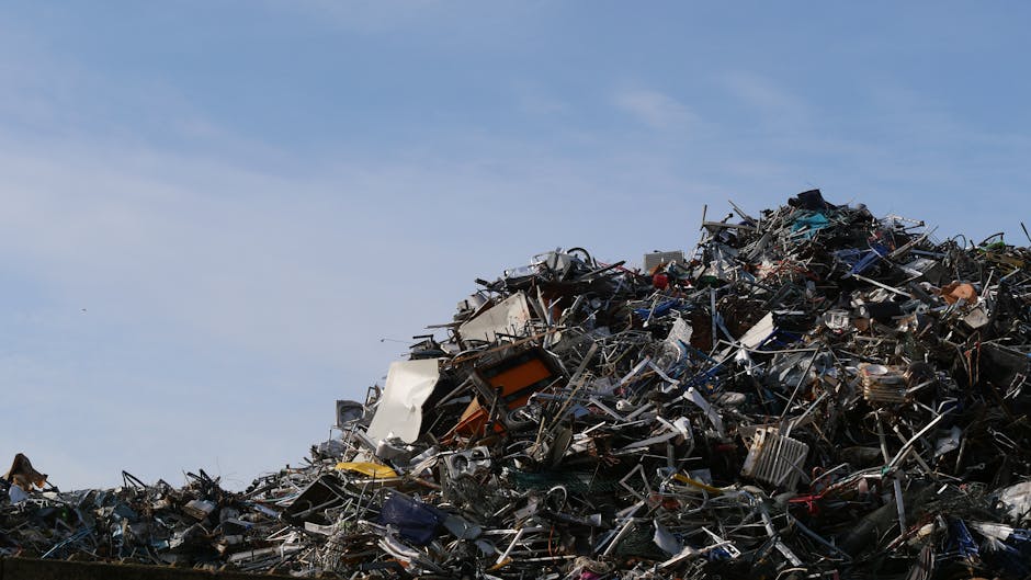 A pile of mixed refuse including black plastic rubbish bags, a yellow plastic container, and a large worn beige car tyre positioned on its side, all situated on an asphalt ground in an open outdoor ar
