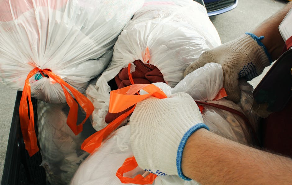 A person wearing a bright orange coat, matching orange hat with a white face mask, and white gloves is seen cleaning under a concrete bridge or overpass. They are using a long-handled brush to remove