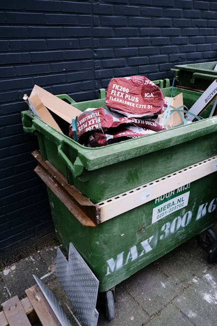 An open green plastic waste bin positioned on a grey concrete pavement next to a dark brick wall, filled with several red and purple bags of construction or building materials, including bags labeled 